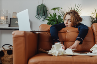 woman looking burnt out and tired slumping over a couch with a laptop and crumbled papers scattered around
