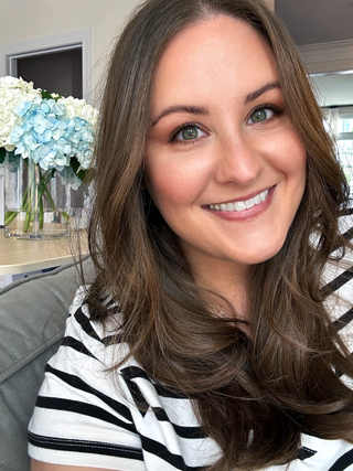 A smiling woman with long brown hair wearing a black and white striped shirt, photographed in a bright indoor setting with flowers and a window in the background.